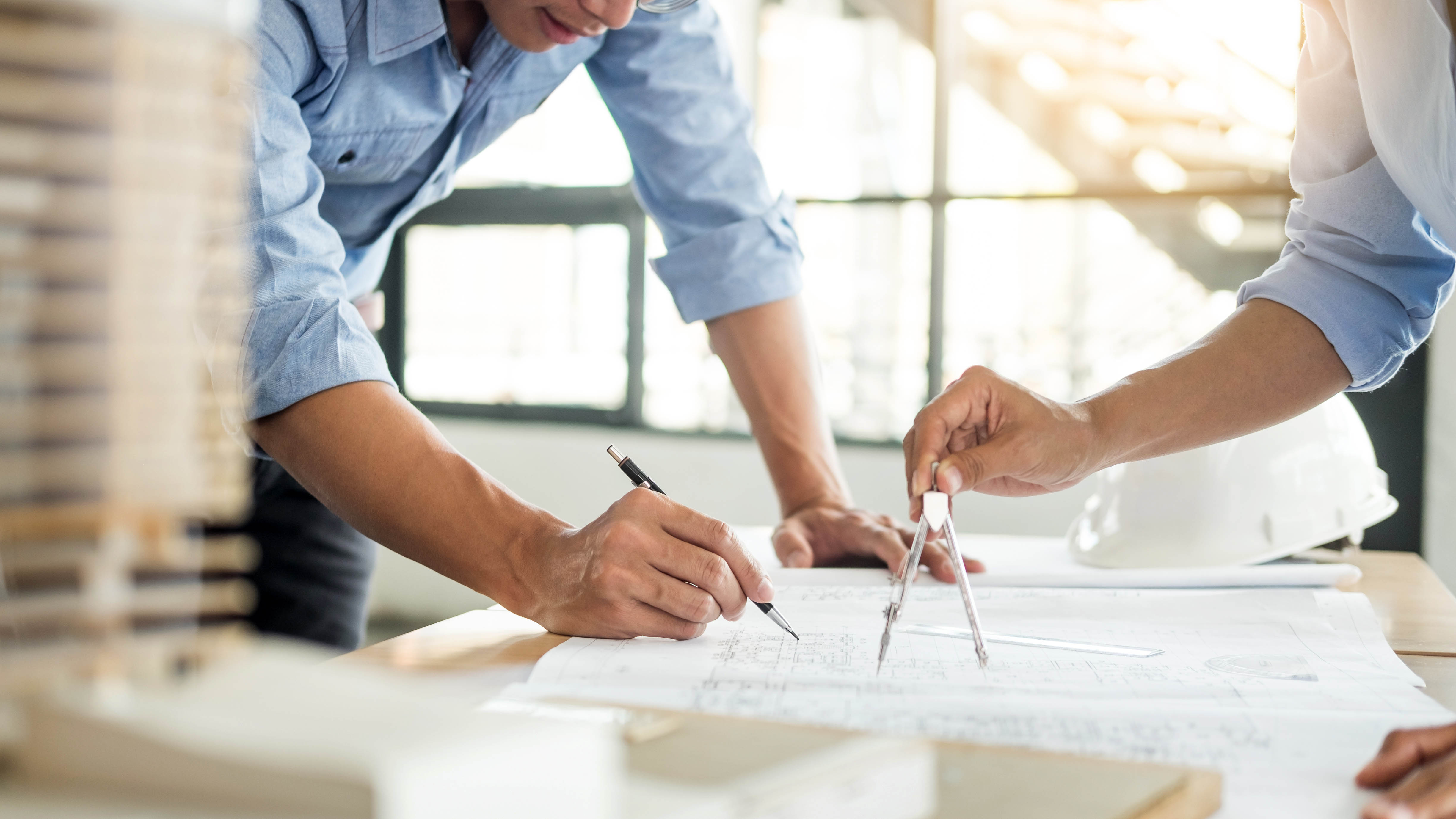 Close-up on torsos, arms, and hands of two professionals working on a blueprint in an office setting.